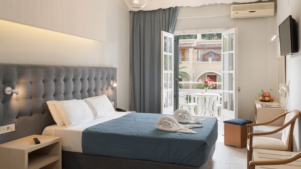 A hotel room with a large bed, a bedside table, a chair, a TV, and a balcony with a table and chairs. - Alkion Hotel Sidari (Corfu)