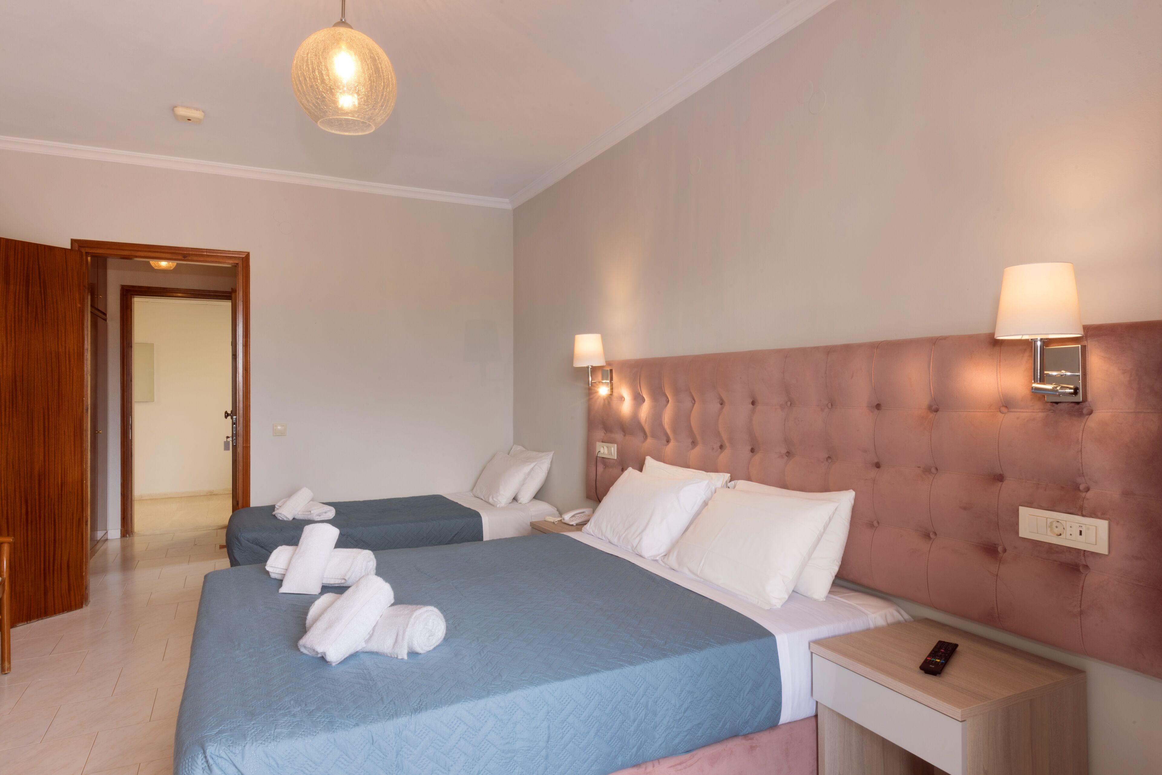 A hotel room with two beds, a wooden door, a bedside table with a lamp, and a wall-mounted light fixture.