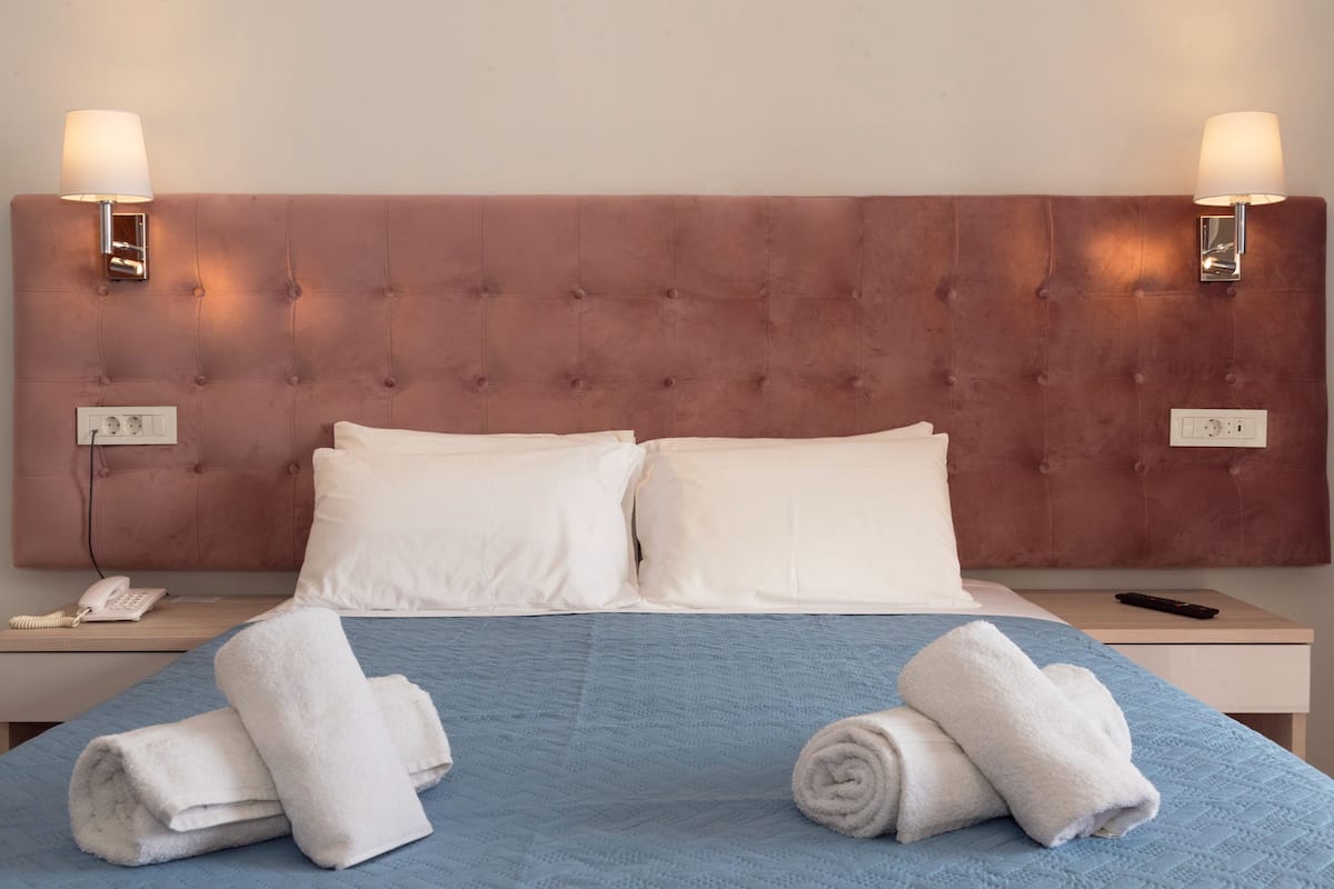 A neatly made bed with white pillows and a blue bedspread, two bedside lamps, a phone, and a remote control on a wooden nightstand.