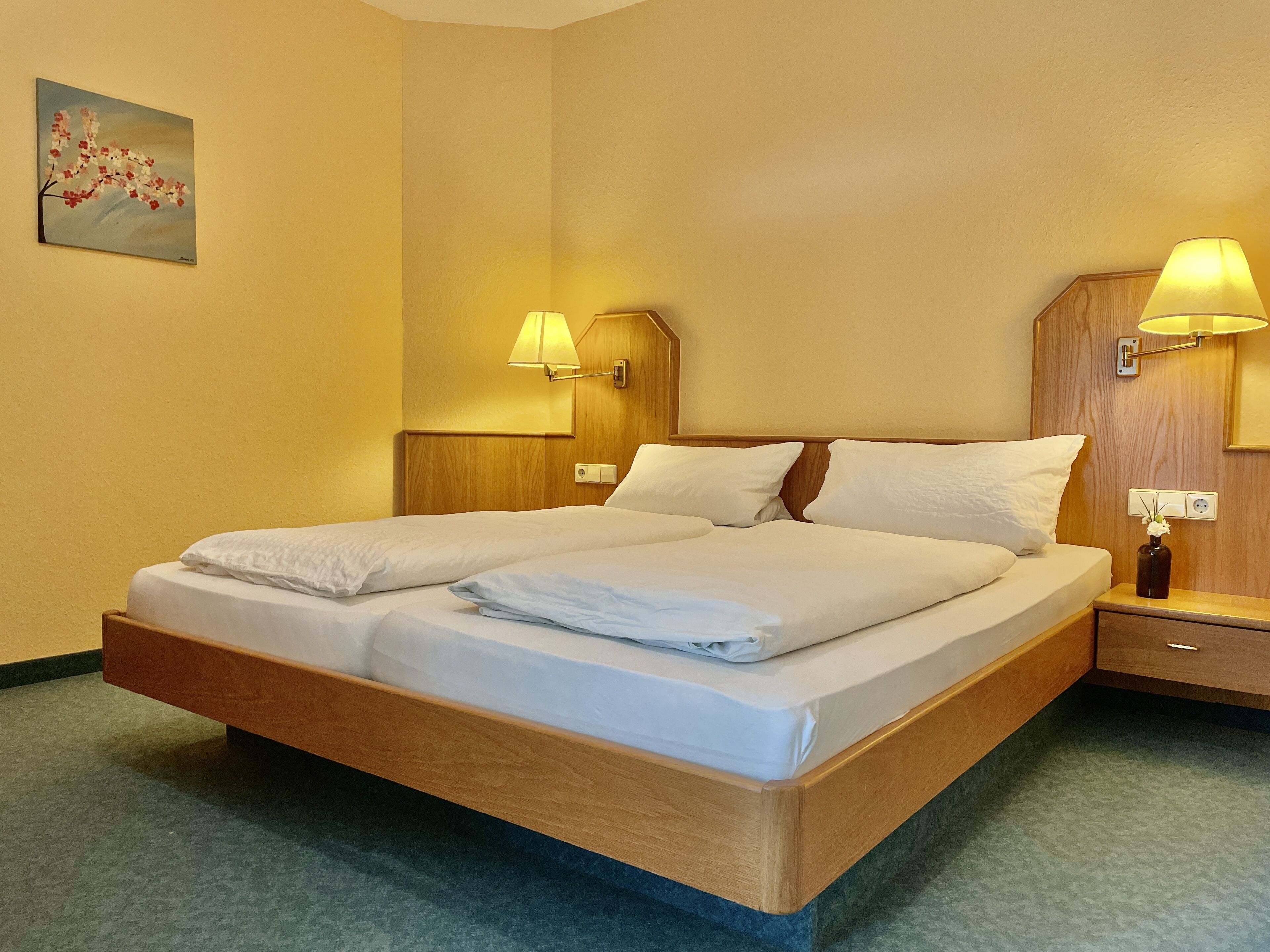 Quarto de hotel com cama de madeira, duas luminárias de cabeceira, mesa de apoio e um quadro na parede.