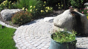 A garden pathway with a stone pavement, large rocks, and various plants including yellow flowers and green foliage. - Landhotel zur Linde (Lossburg)