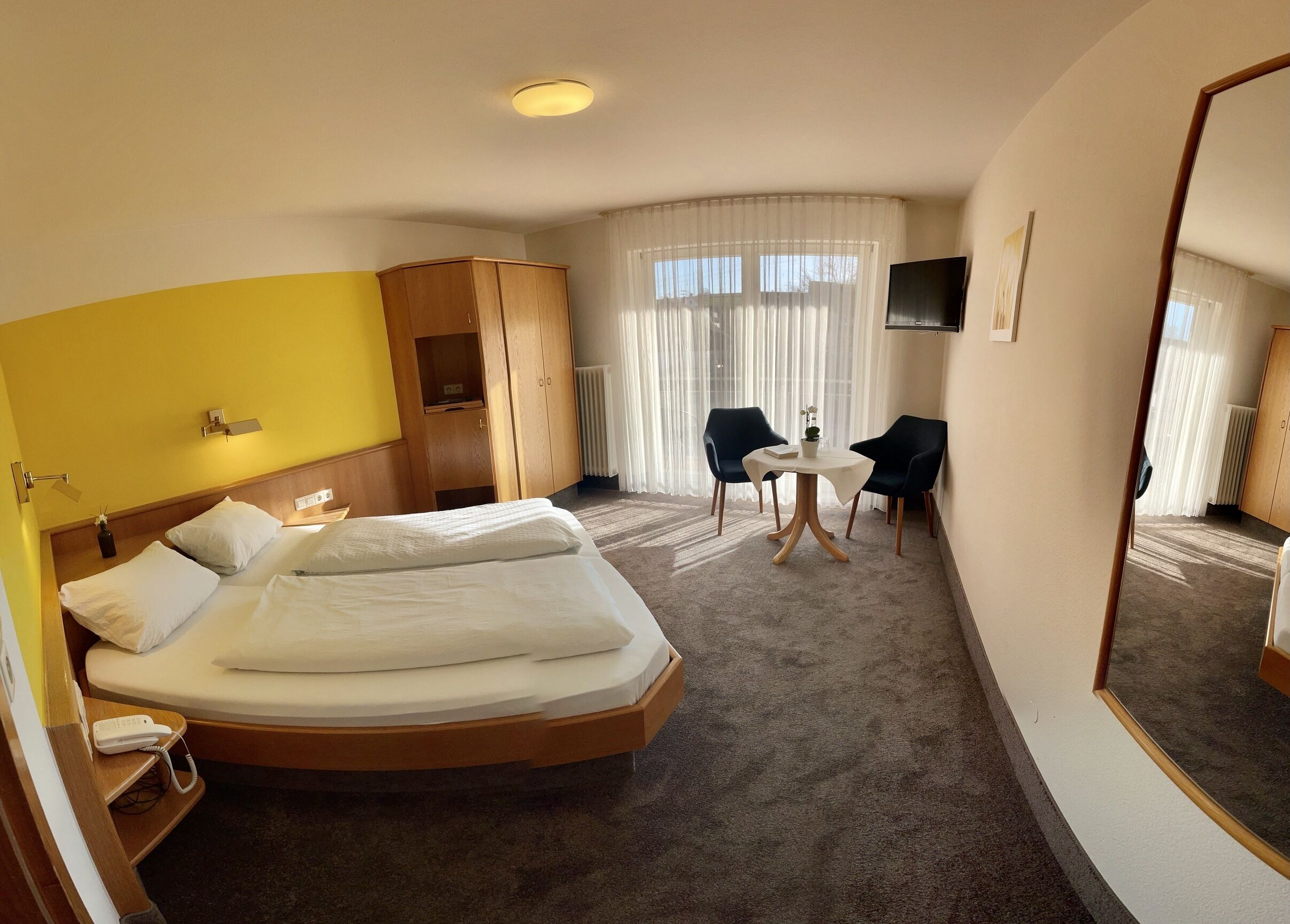 Een hotelkamer met een groot bed, een houten garderobe, een kleintje tafel met twee stoelen, een televisie en een grote spiegel.