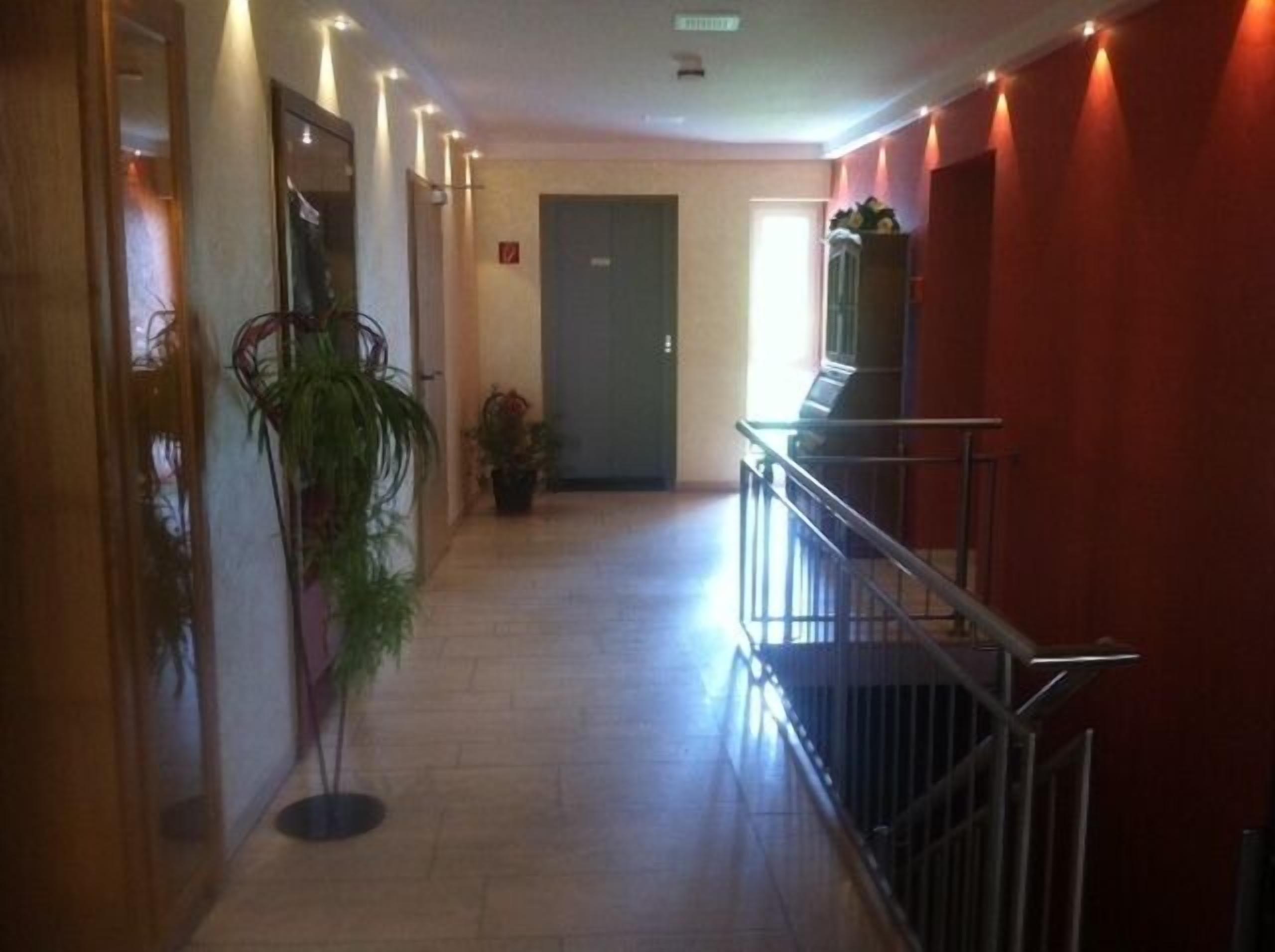 A hotel corridor with a wooden floor, a potted plant, a door at the end, and a railing on the right.