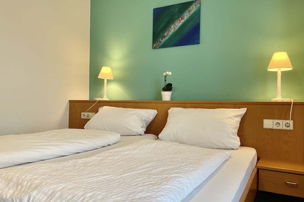 A double bed with white bedding, two bedside lamps, a small potted plant, and a painting on the wall.