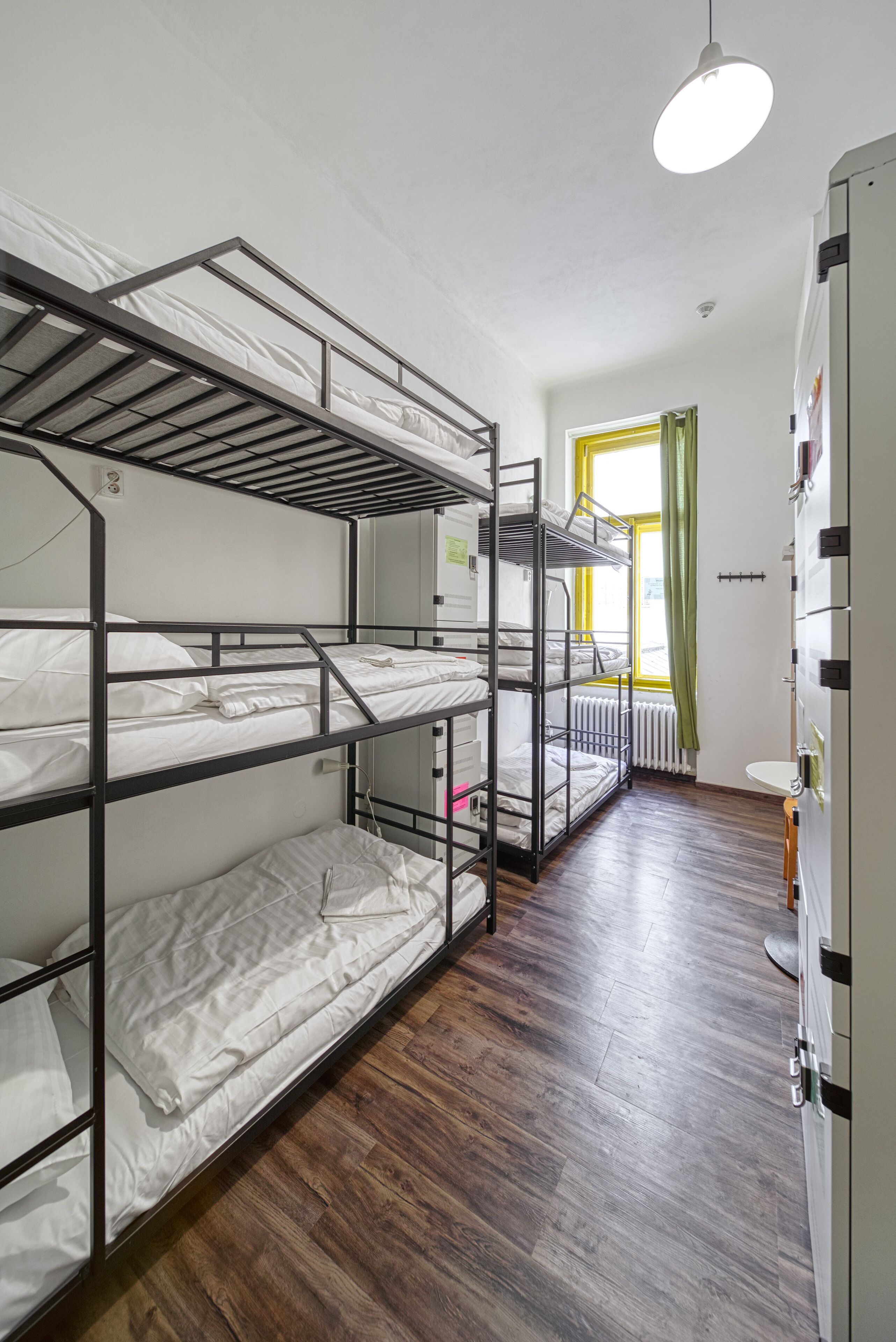 A dormitory room with bunk beds, a radiator, a window with curtains, and a sink.