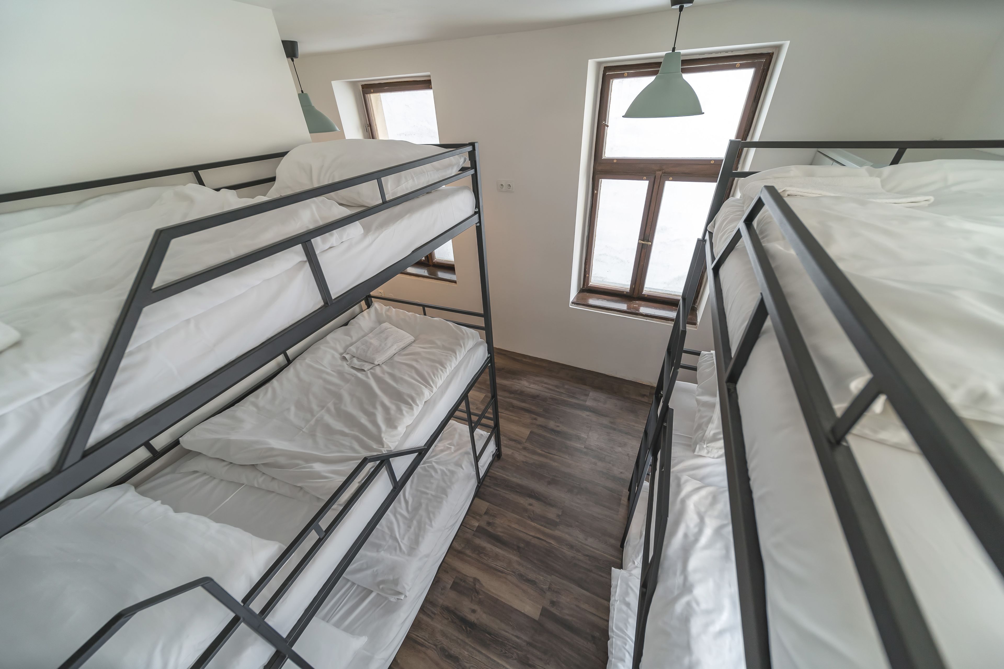 A dormitory room with bunk beds, wooden flooring, and windows with curtains.