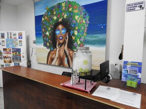 A reception desk with a large painting of a woman surrounded by flowers, a jar of lemons, and various informational posters. - Hostal Don Miguel Cartagena de Indias (Cartagena)