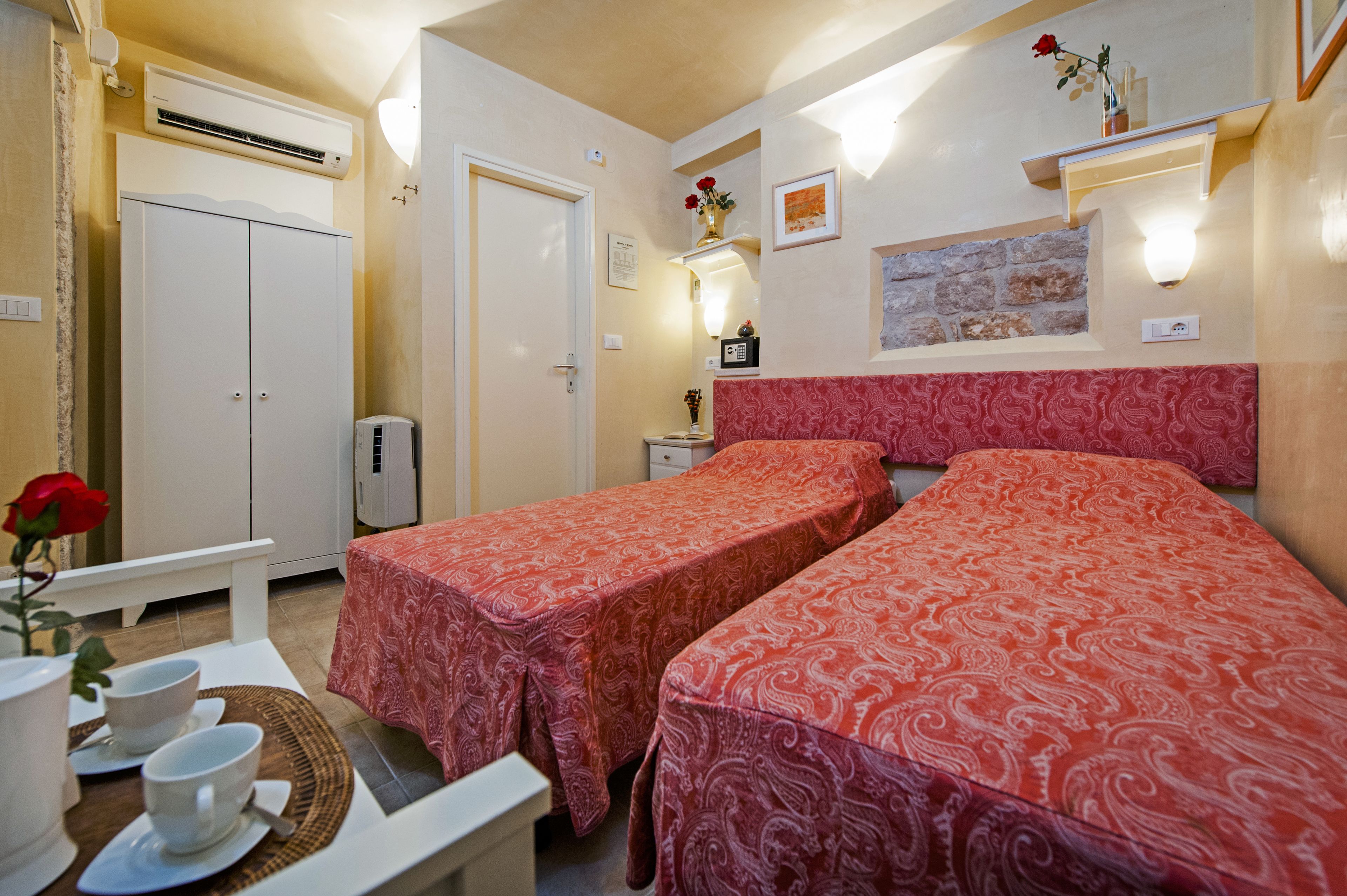 A hotel room with two beds, a desk with a chair, a small table with cups and a vase of flowers, a wardrobe, an air conditioning unit, and a wall-mounted shelf with a vase of flowers.