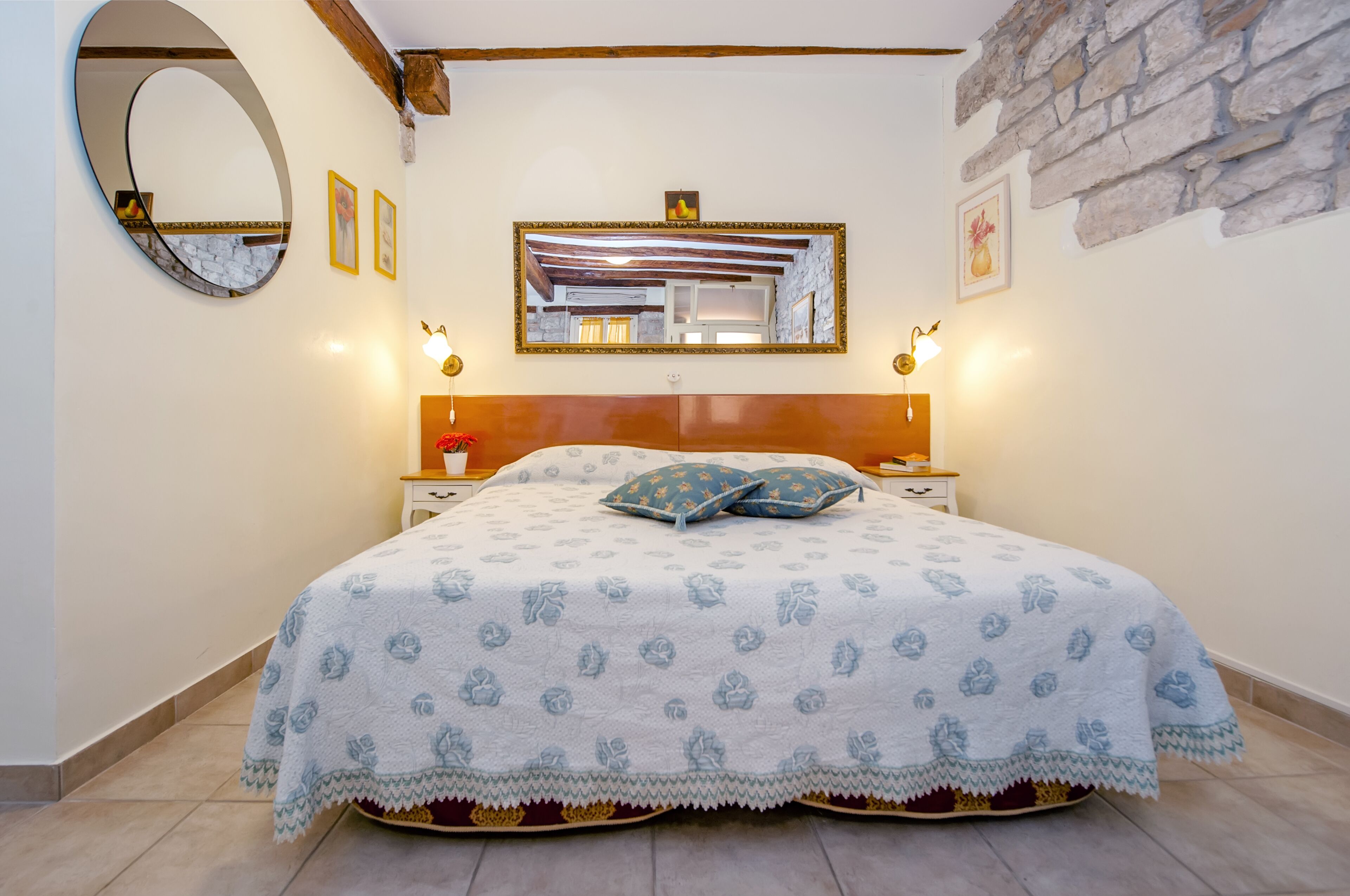 A bedroom with a bed, a round mirror, a wooden headboard, a stone wall, and a framed picture.