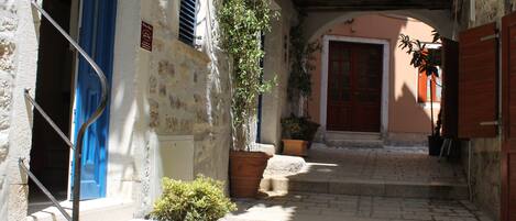 A narrow alleyway with stone steps leading to a building with a red door. There are potted plants along the path and a metal railing on the left.