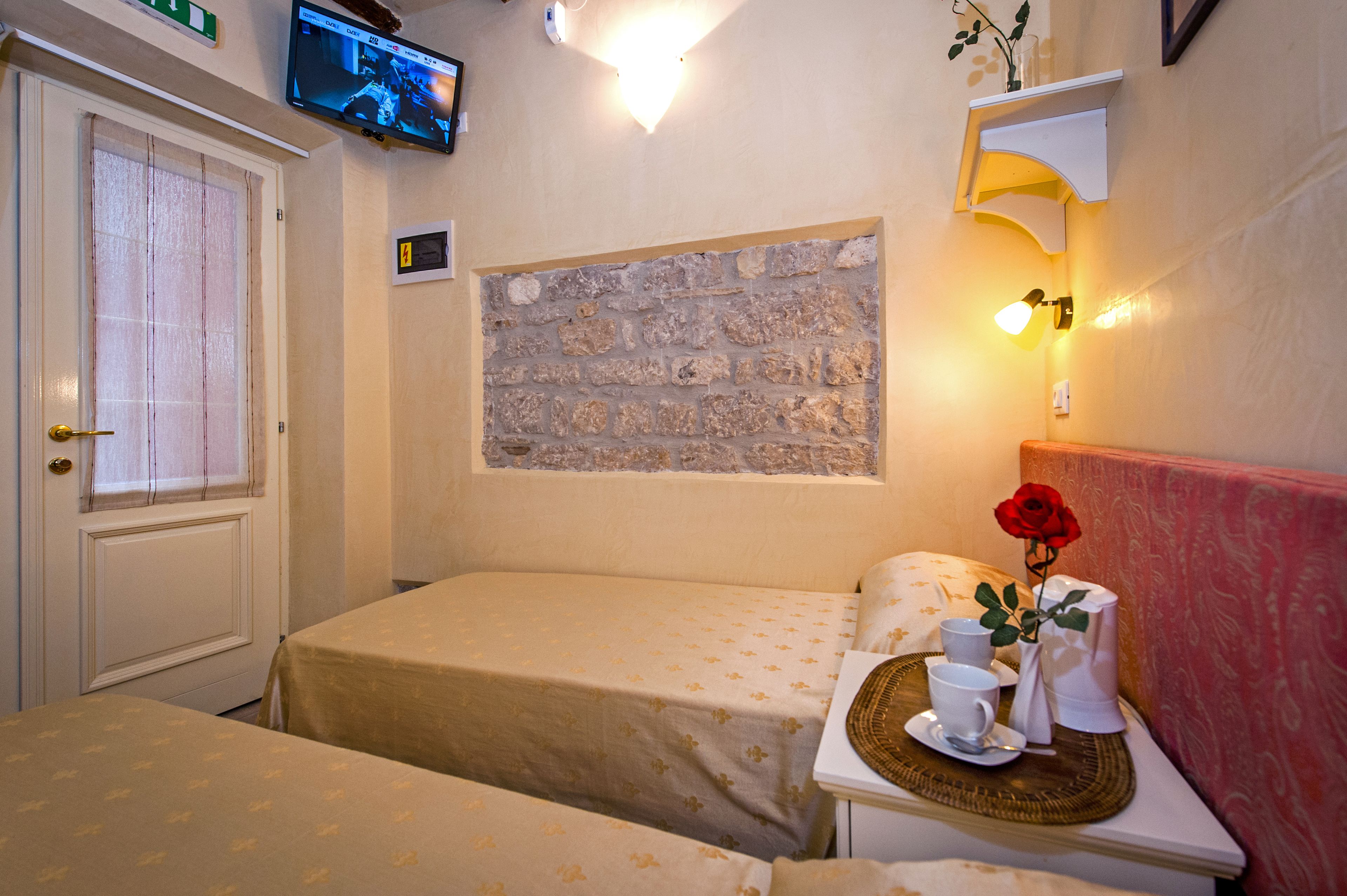 Habitación de hotel con dos camas, un televisor en la pared, un muro de piedra y una mesita de noche con un jarrón de una rosa roja.