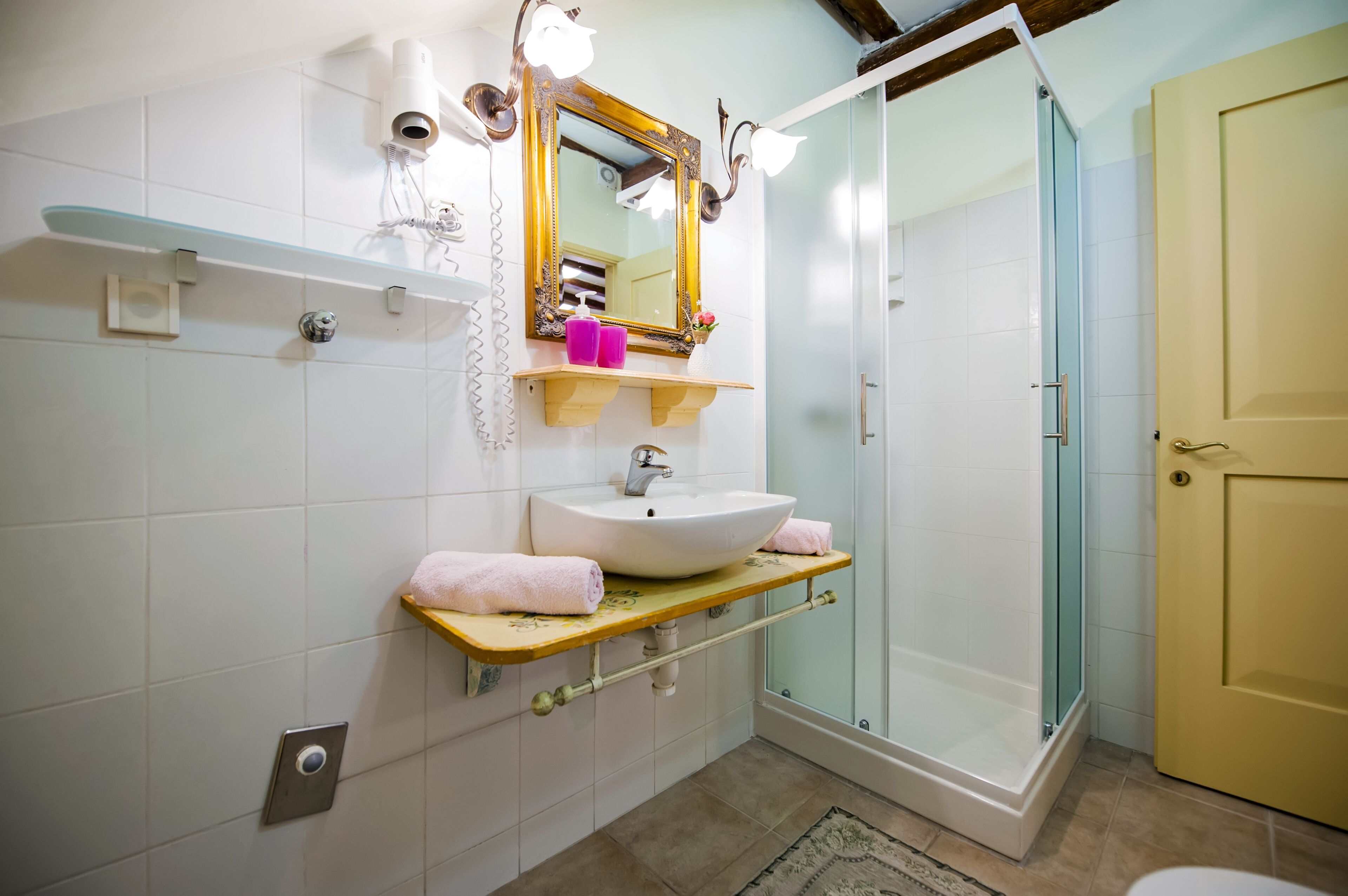 Un baño con cabina de ducha de vidrio, un lavabo blanco con encimera de madera, un espejo con marco dorado y una puerta amarilla.