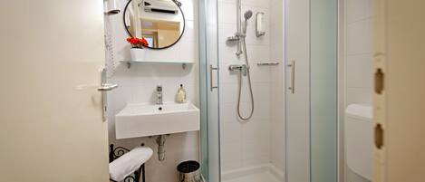 Un baño con ducha envuelta en vidrio, un lavamanos blanco, un espejo con marco decorativo y un pequeño cubo de basura.