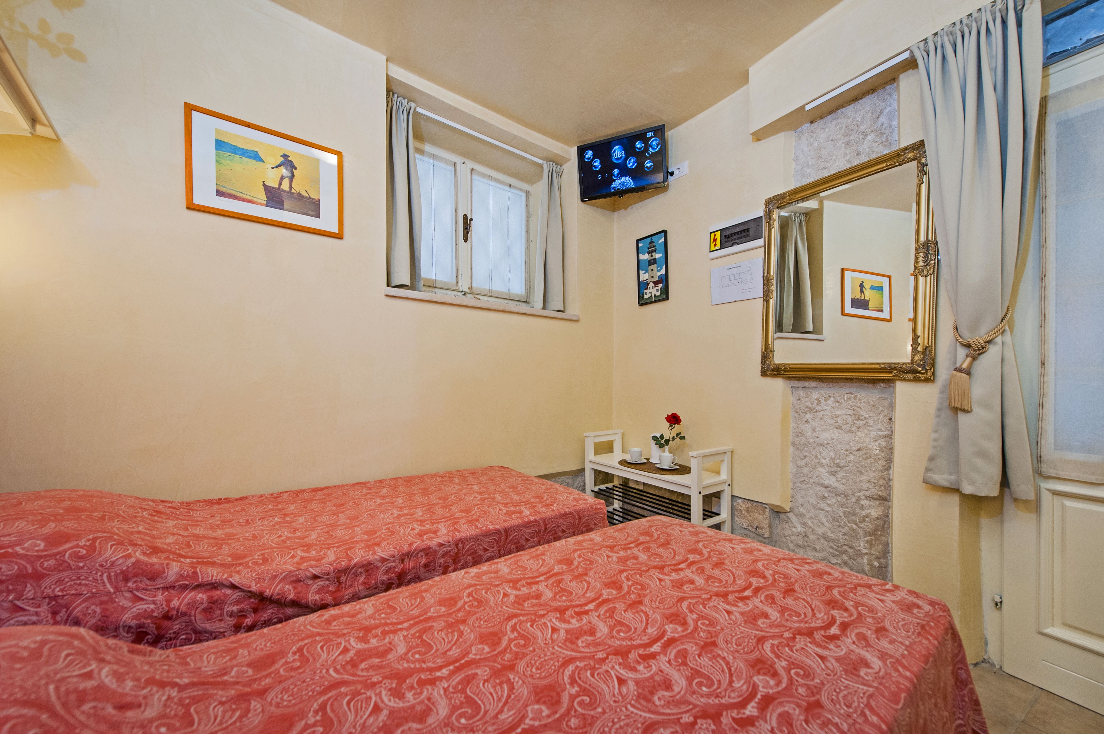 Una habitación con dos camas, televisión, ventana con cortinas, espejo y una mesita con un florero.