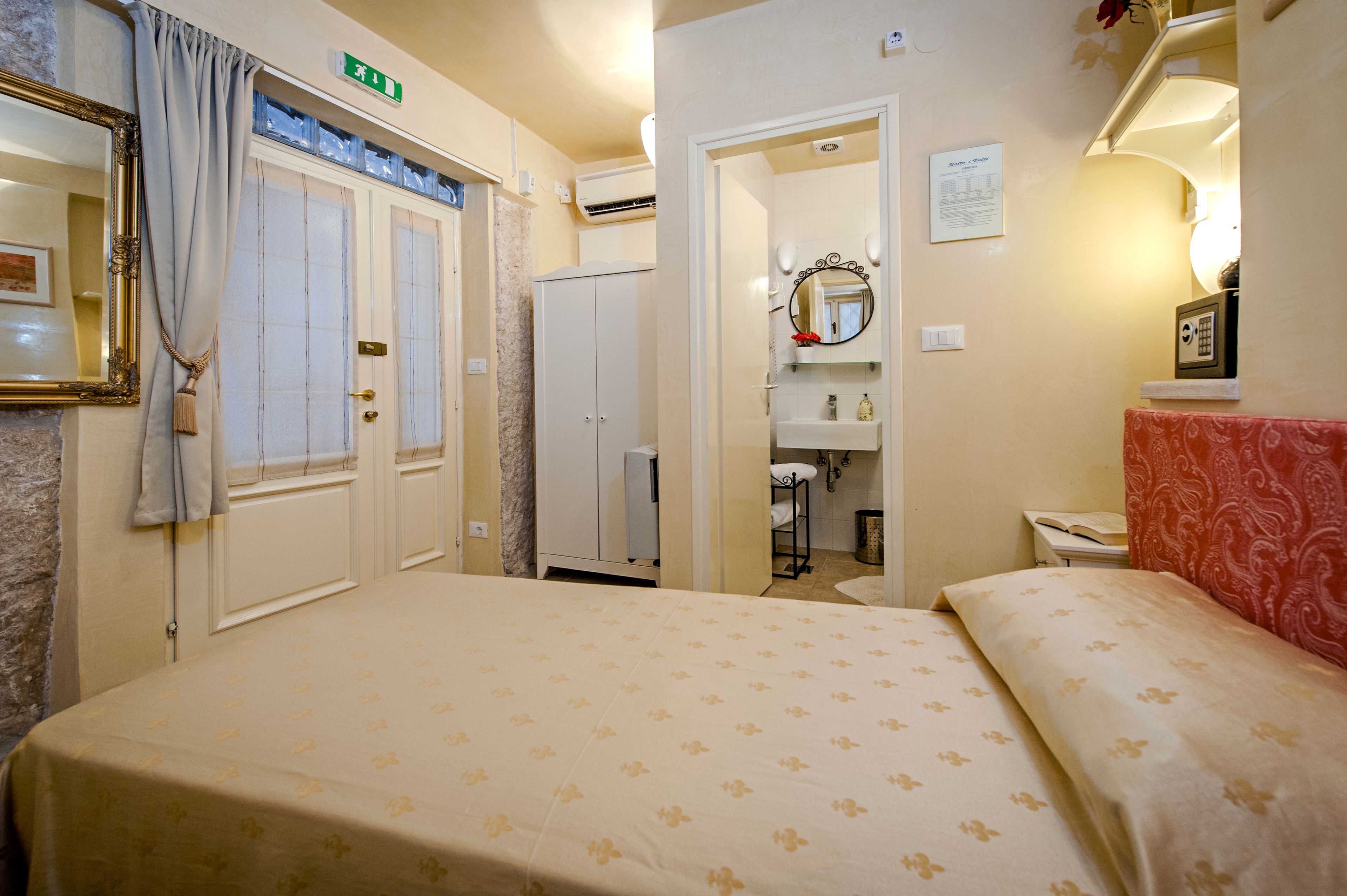 A hotel room with a bed, a bathroom with a mirror, and a red patterned headboard.