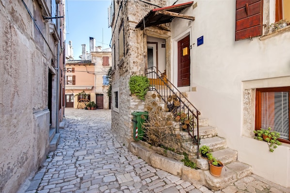 Una estrecha calle adoquinada bordeada por edificios históricos con fachadas de piedra, una puerta de madera y una pequeña escalera que conduce a un segundo piso.