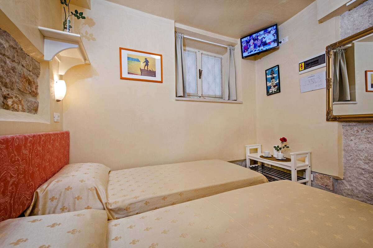 Ein Hotelzimmer mit zwei Betten, einem Fernseher, einem kleinen Tisch mit Blumenvase und einem Fenster mit Vorhängen.