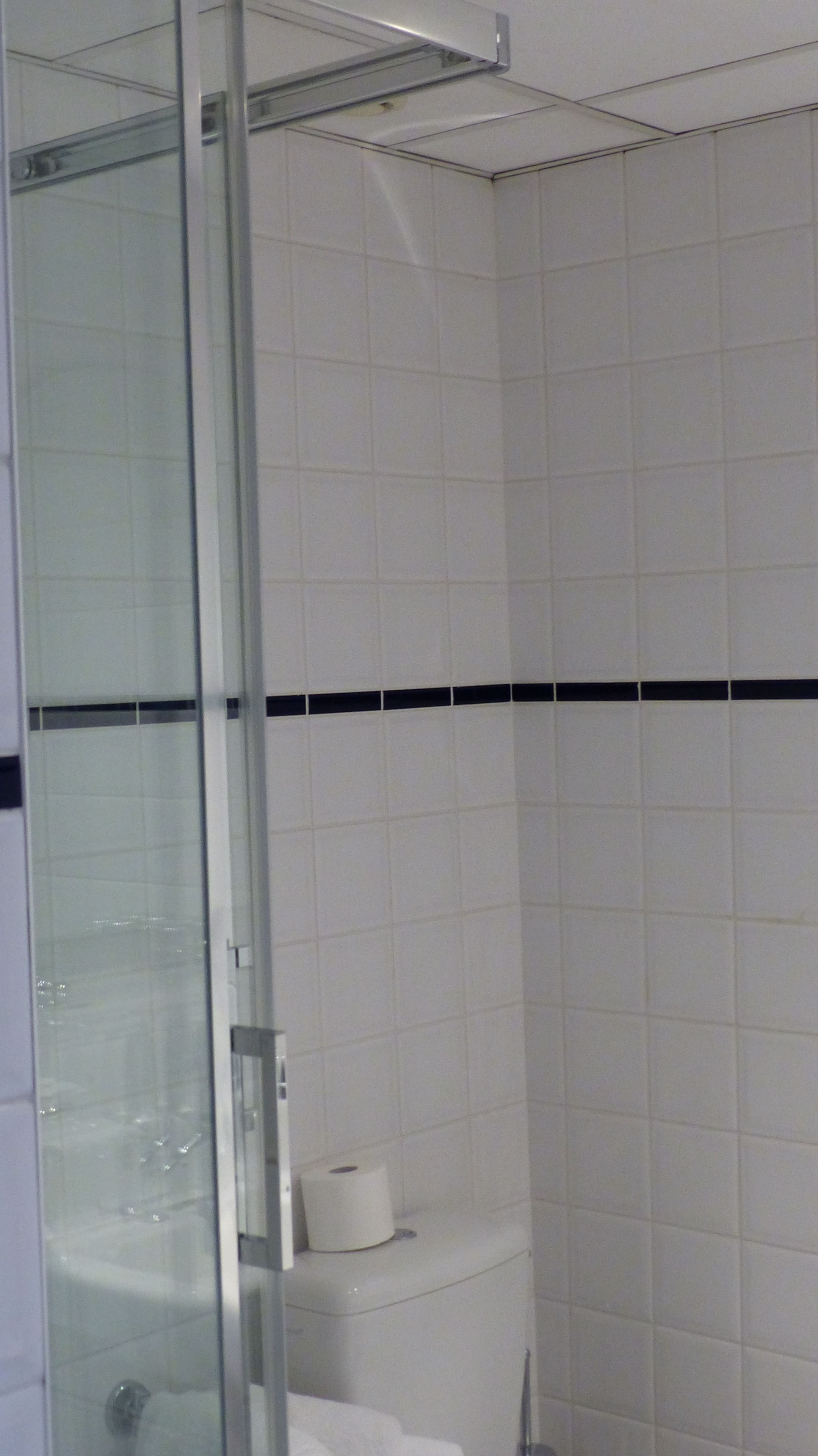 A bathroom with white tiled walls, a glass shower enclosure, and a toilet with a roll of toilet paper on top.