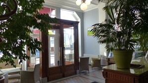 A lobby with wicker furniture, a large potted plant, and a wooden door leading outside. - Hôtel de l'Europe (Toul)