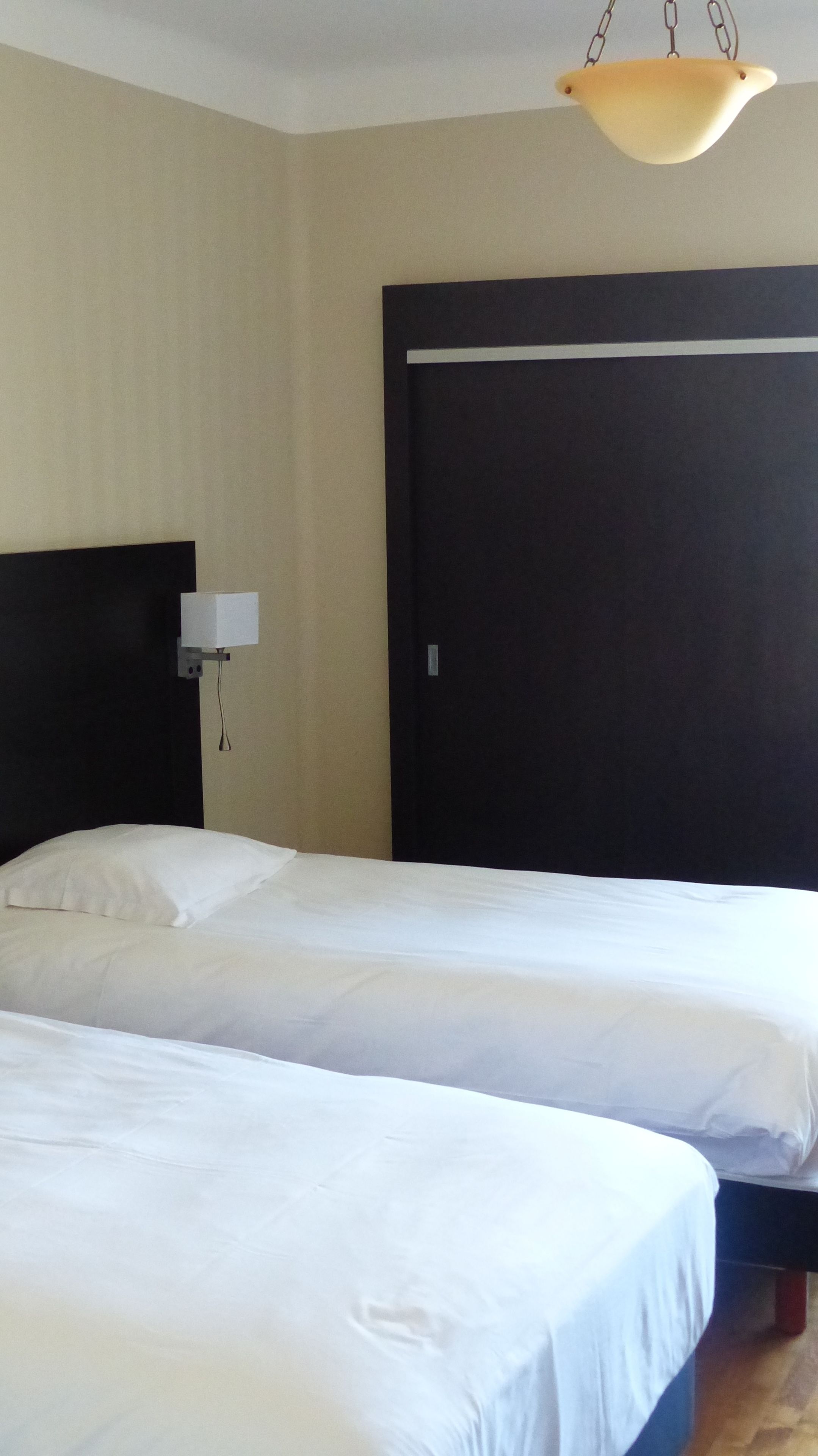 A hotel room with two beds, a dark headboard, a wall-mounted lamp, and a hanging light fixture.
