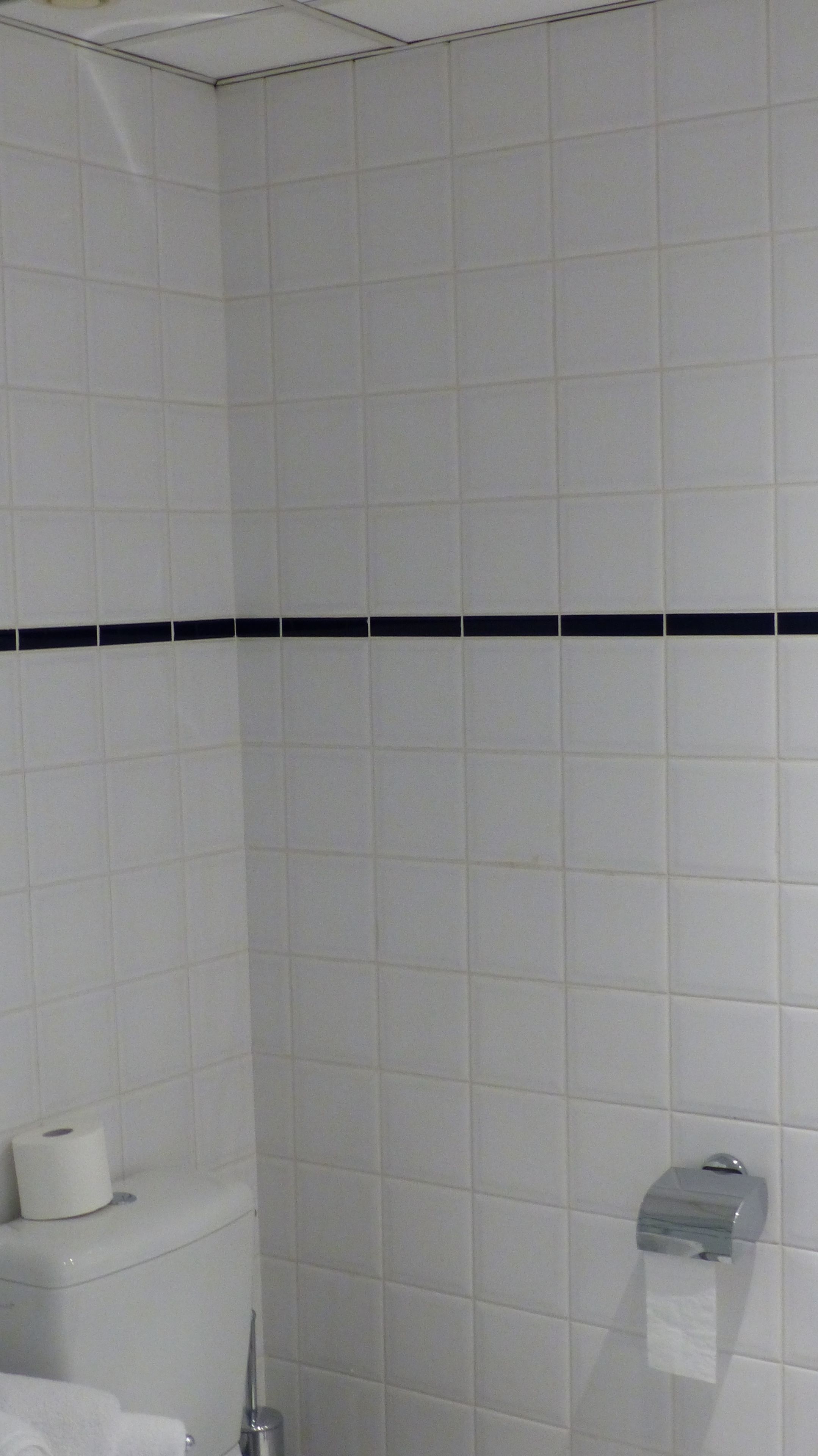 Un baño con paredes de azulejos blancos, una banda horizontal negra, un inodoro y un dispensador de toallas de papel.