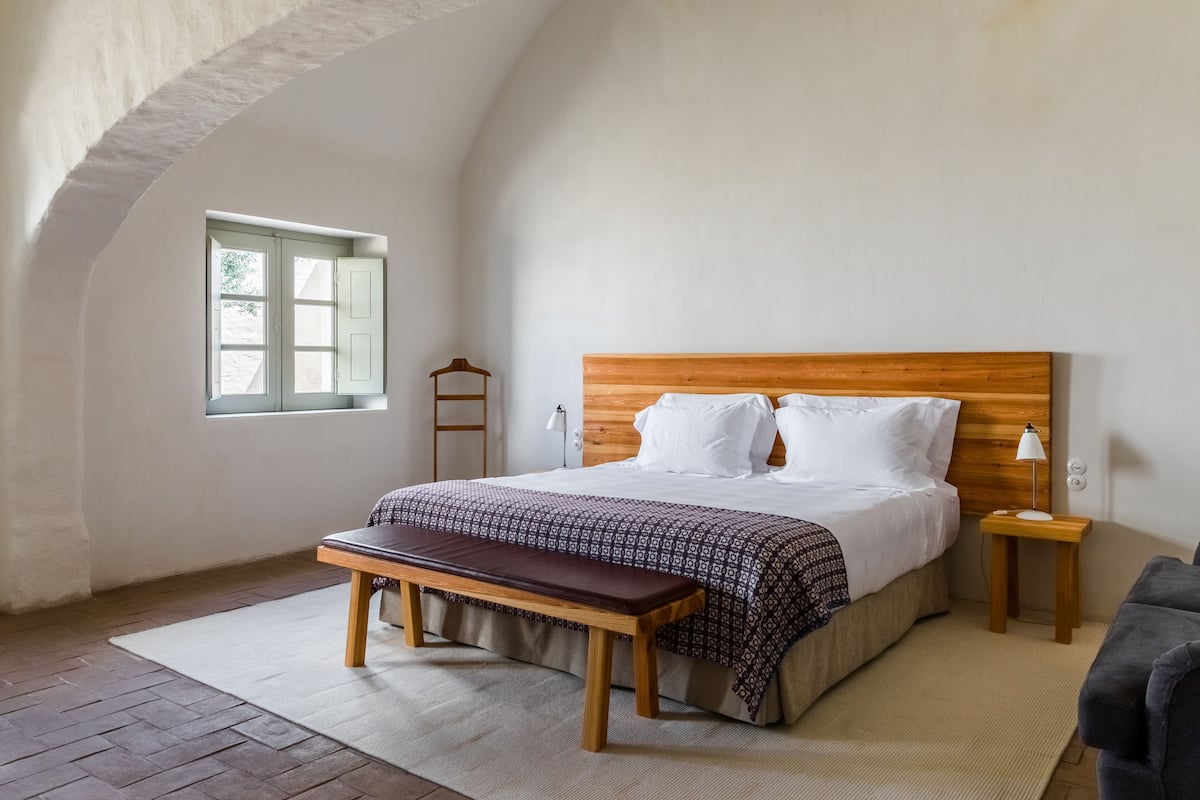 Ein Schlafzimmer mit einem Holz-Kopfteil, einem Bett mit weißer Bettwäsche, einer Bank, einem Nachttisch mit einer Lampe und einem Fenster mit Holzrahmen.
