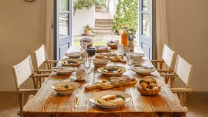 Une table de salle à manger en bois, dressée avec des assiettes, des bols, des verres et des couverts, donnant sur un jardin par une porte ouverte.