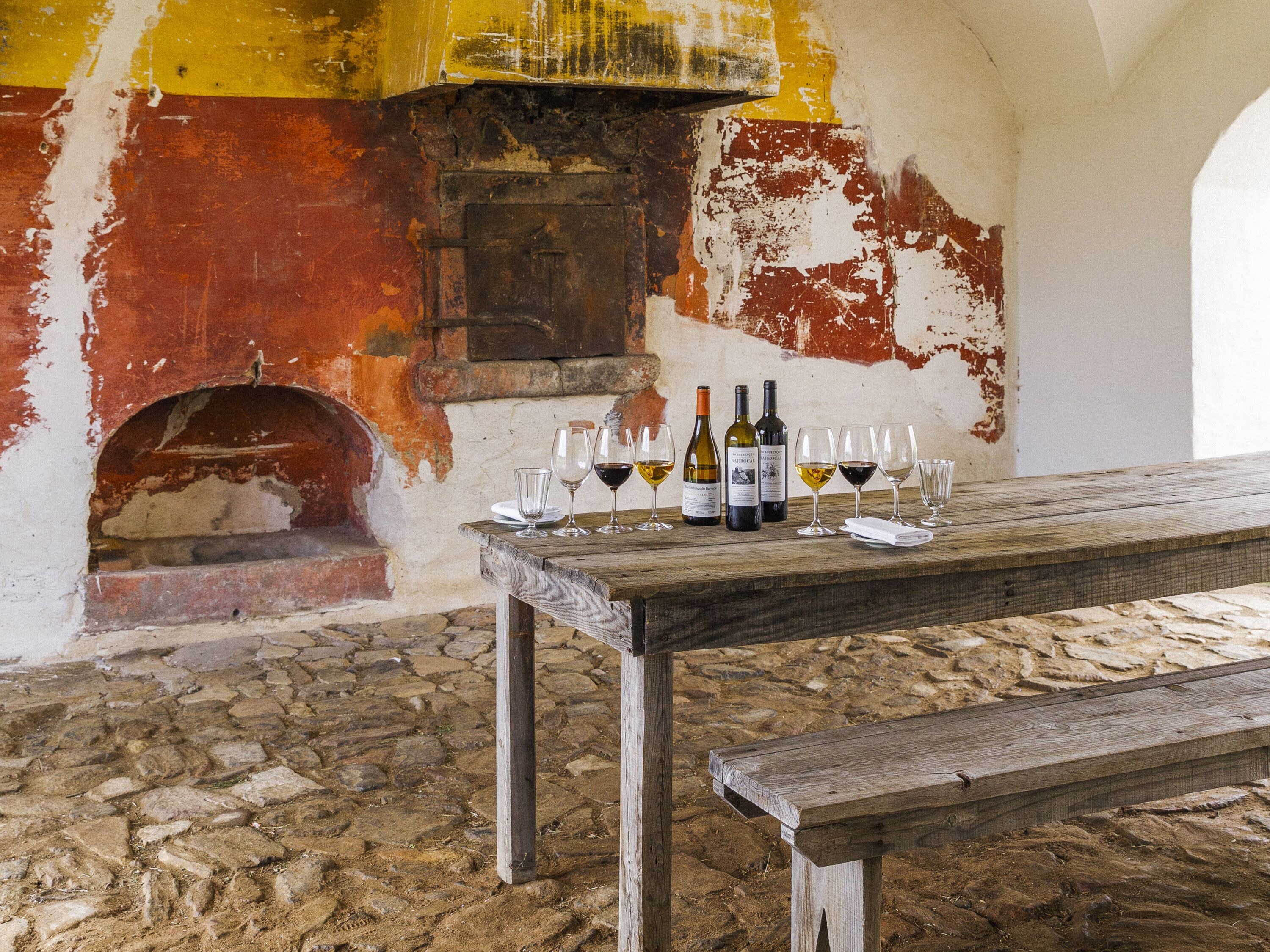 A rustic interior with a wooden table displaying wine bottles and glasses, a stone floor, and a weathered wall with peeling paint.