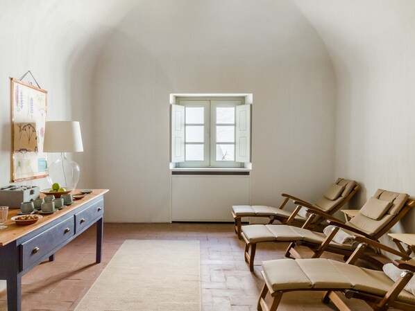 A room with a wooden desk, a lamp, a window with shutters, and several lounge chairs.