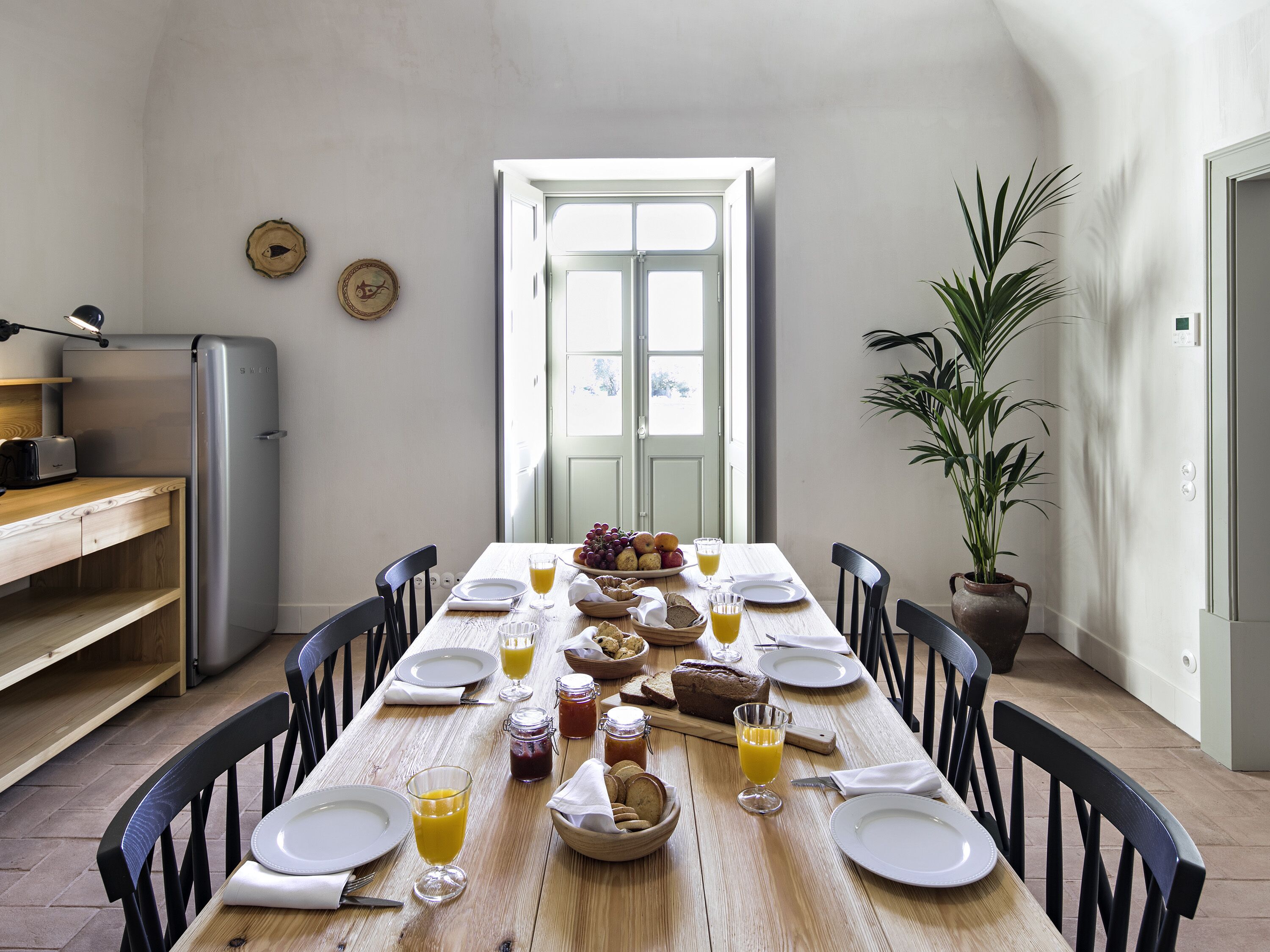 A dining area with a wooden table set for breakfast, a refrigerator, and a potted plant.