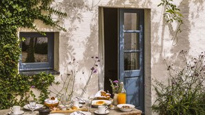 Une table de jardin rustique, dressée à l’extérieur avec différents plats, des verres et une bouteille de jus d’orange, entourée de verdure et d’un bâtiment doté d’une porte et d’une fenêtre bleues.