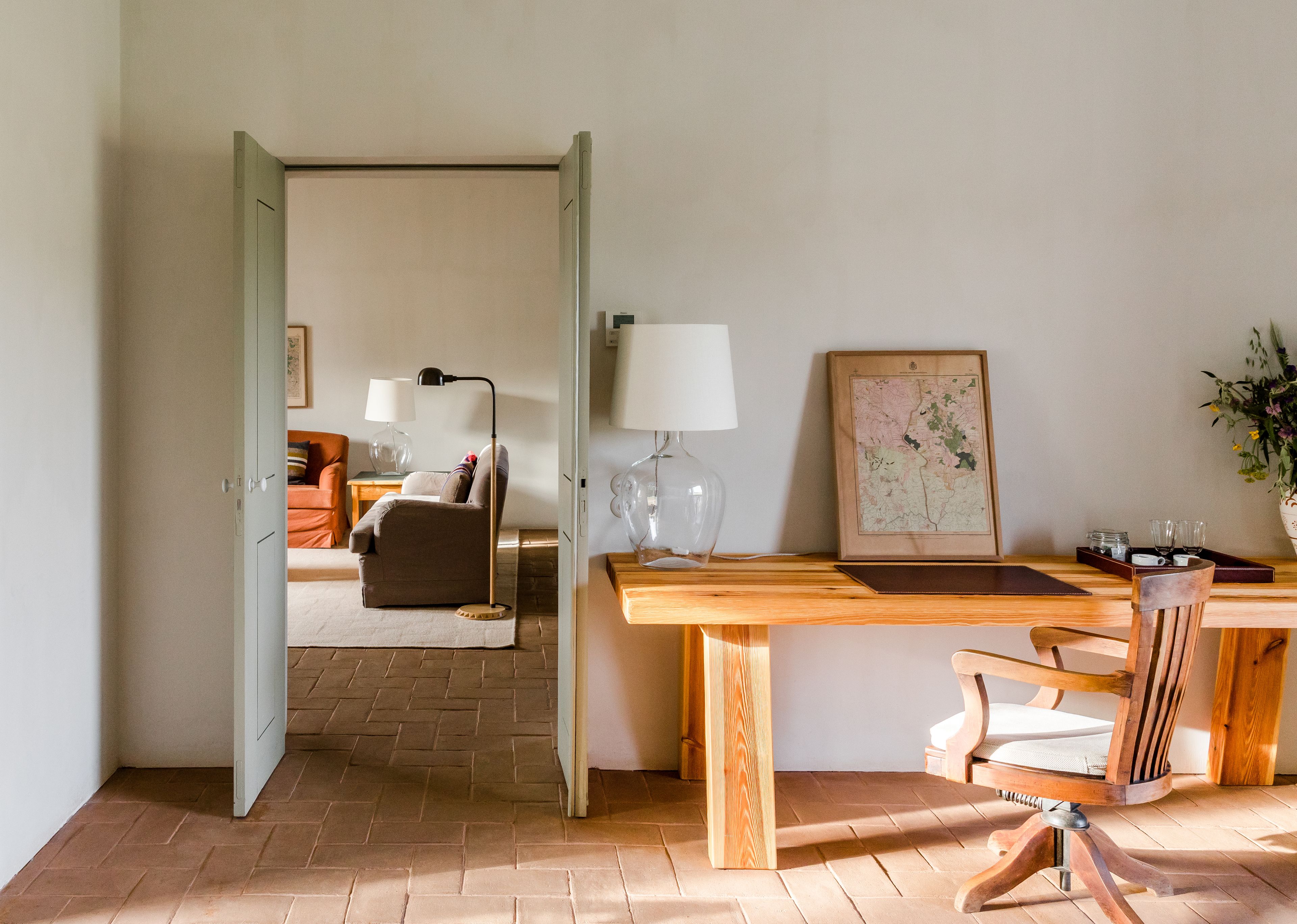 A room with a wooden desk, a chair, a lamp, a map, and a bed with orange bedding.