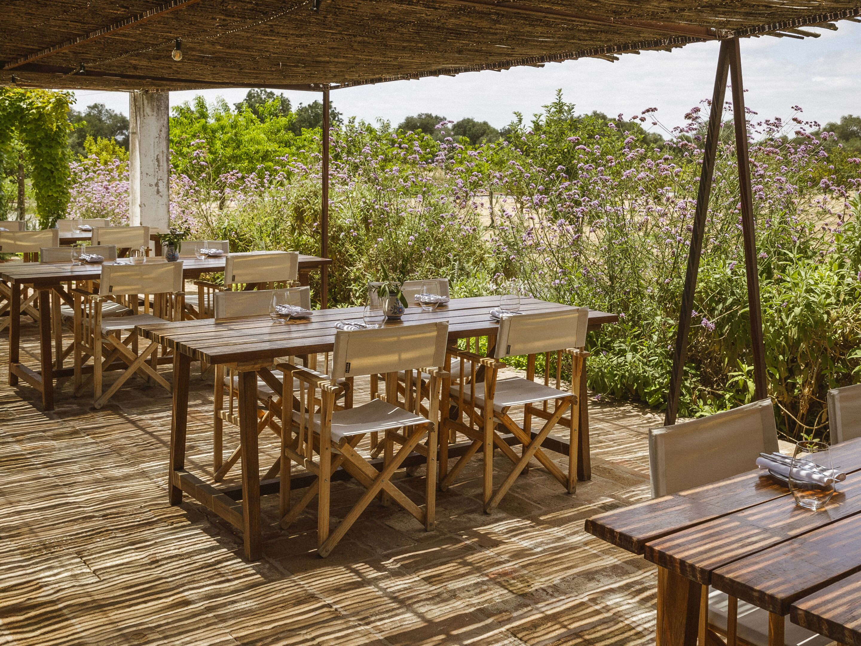 An outdoor dining area with wooden tables and chairs, surrounded by lush greenery and purple flowers.