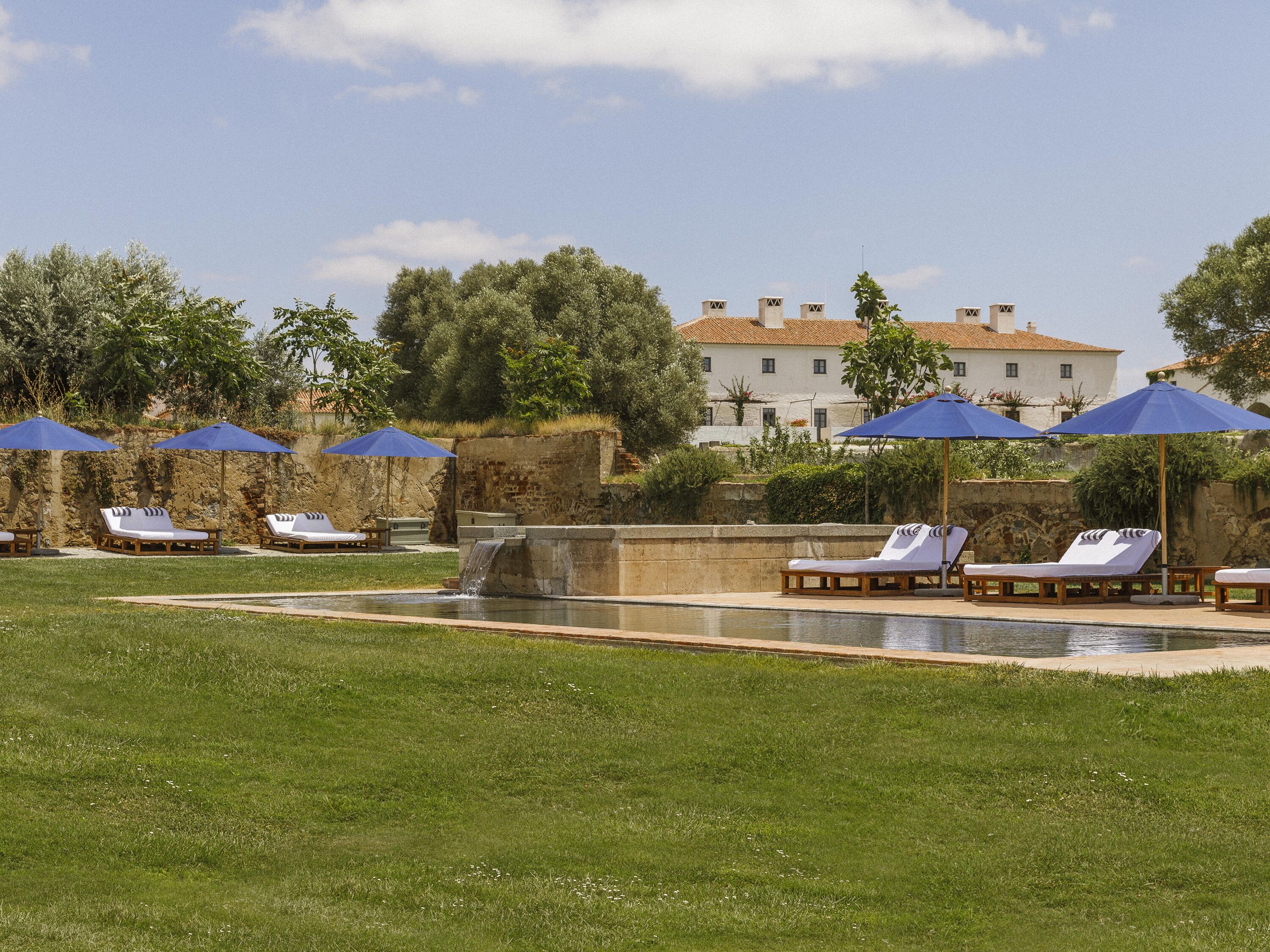 Zona de piscina con sillas de descanso y sombrillas, una pared de piedra y un edificio al fondo.