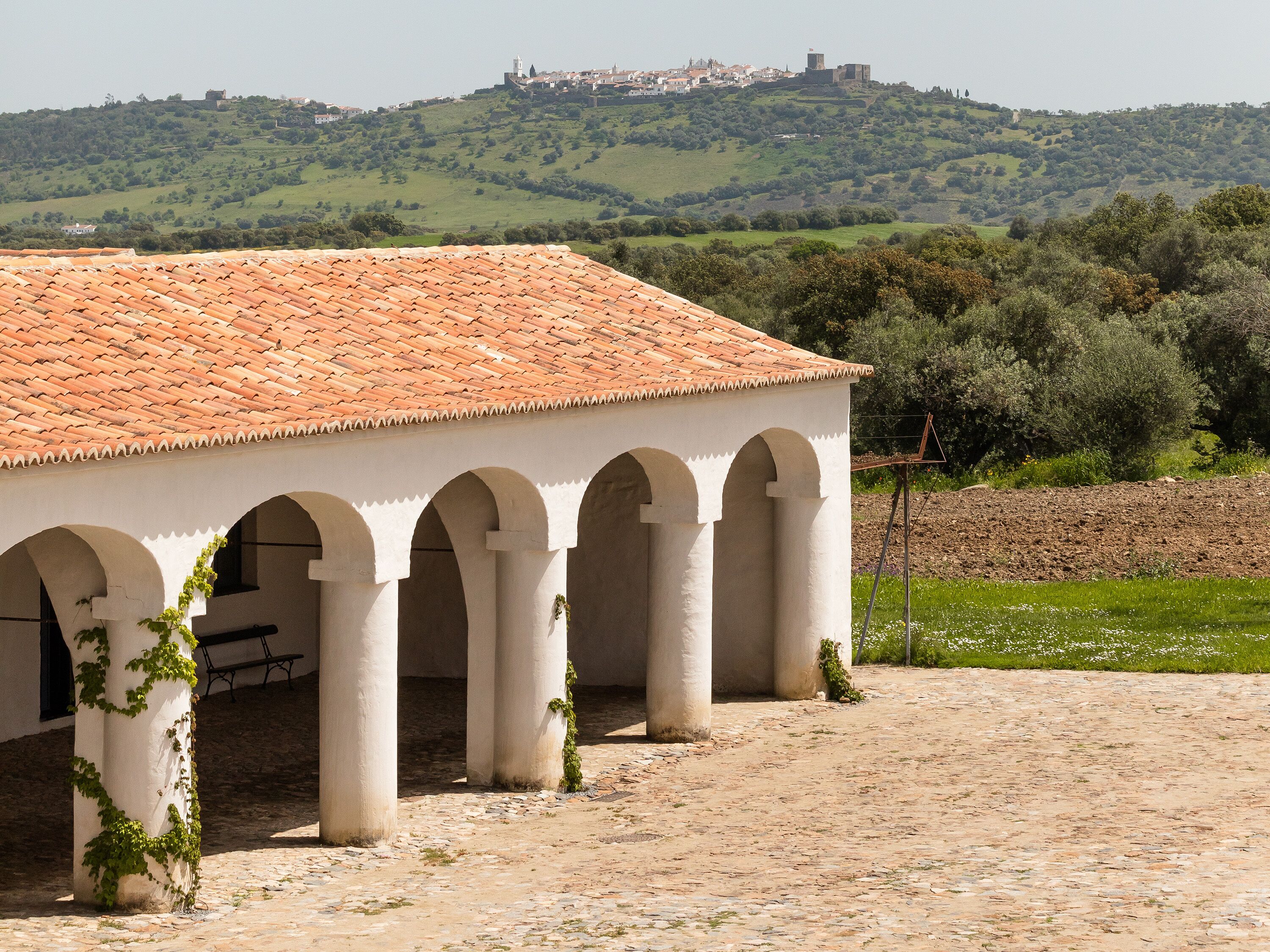A building with a tiled roof and arches, set against a hilly landscape with a castle in the background.