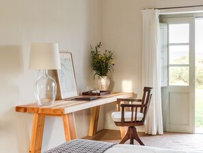 A wooden desk with a lamp, a framed map, a vase with flowers, and a chair in a room with a door leading outside.