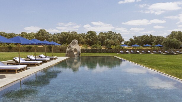 Une piscine entourée de verdure et agrémentée d’une sculpture en pierre, avec des chaises longues et des parasols à proximité.