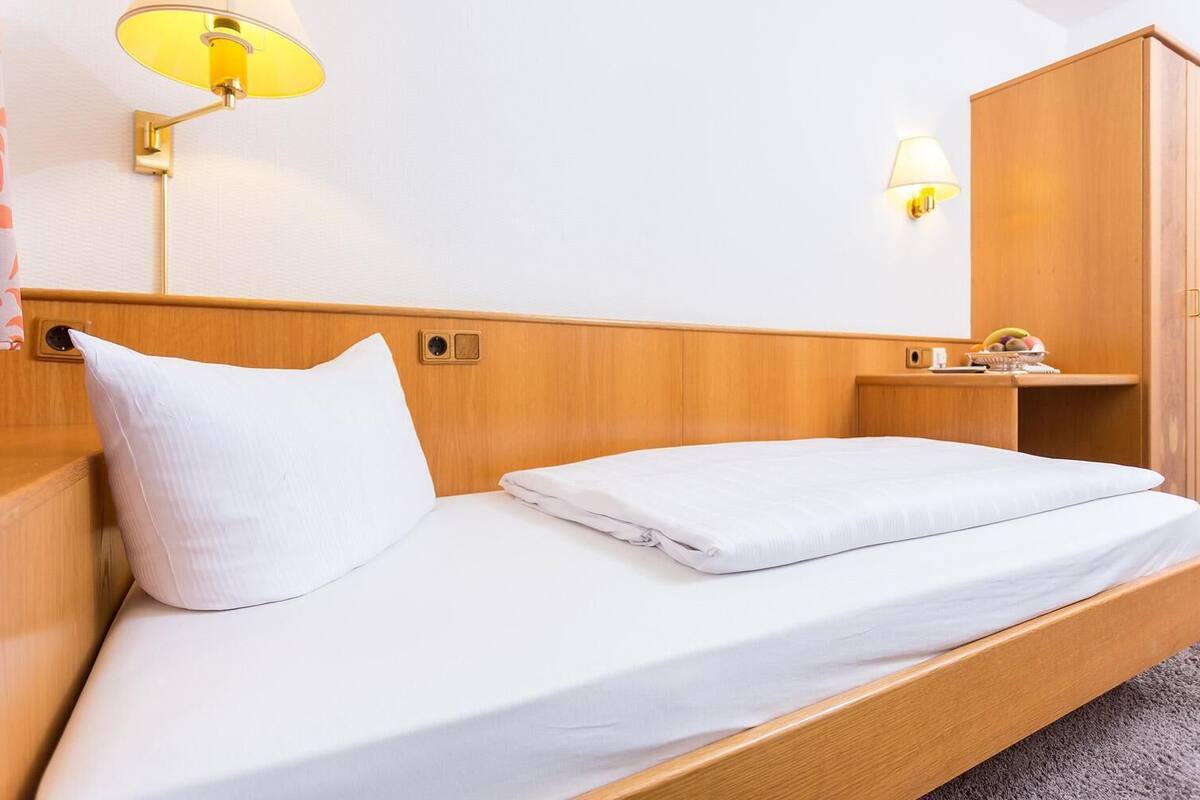 A single bed with a wooden headboard, white bedding, and a nightstand with a lamp.