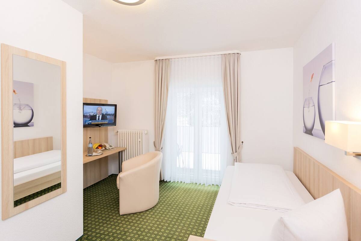 A hotel room with a bed, a desk with a computer, a chair, a television, and a window with curtains.