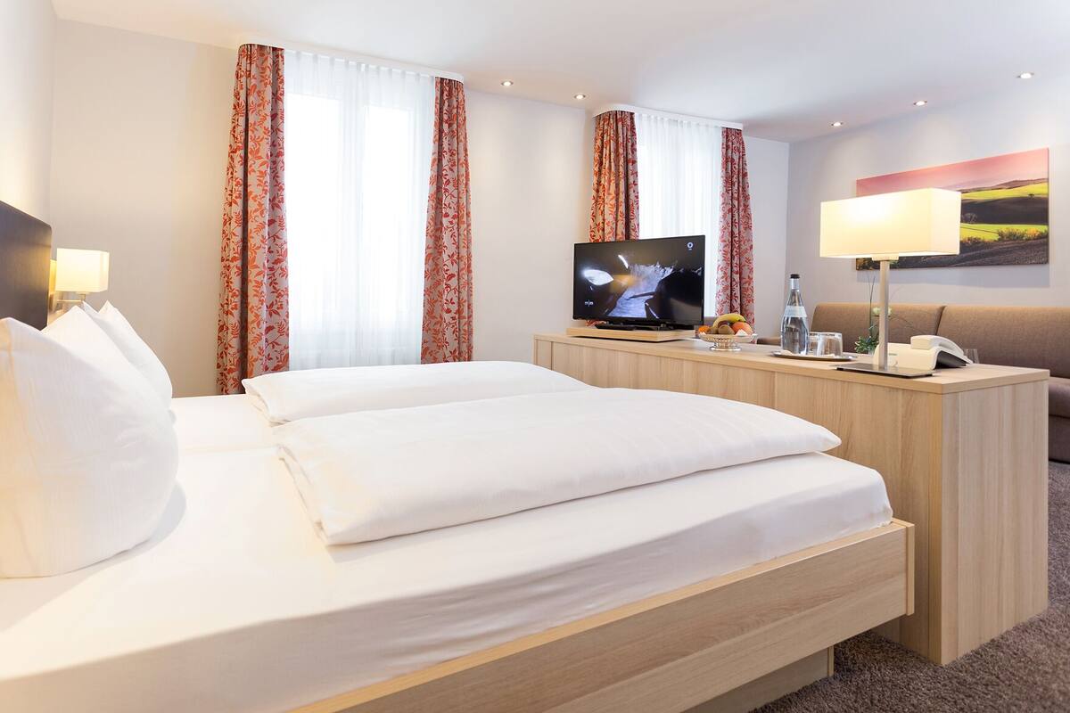 A hotel room with a bed, a TV, a desk with a lamp, and a painting on the wall.