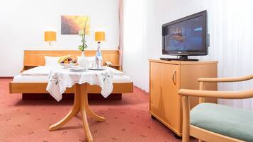 A hotel room with a bed, a wooden nightstand with a television, a table with a vase and fruit, and a chair.