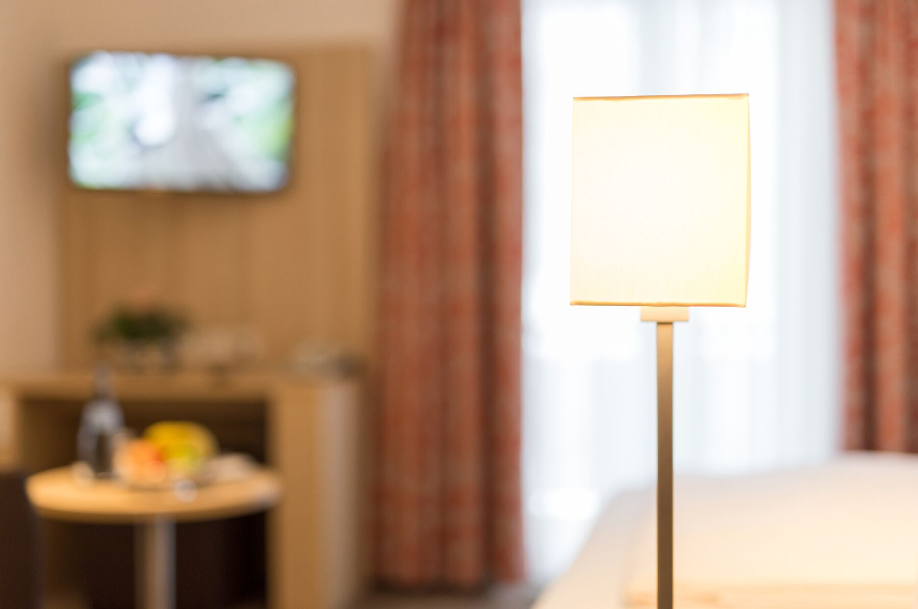 A room with a television on the wall, a table with items on it, and a lamp with a blank screen.