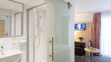 Baño de hotel con cabina de ducha de vidrio, un lavabo y un espejo. Hay un televisor montado en la pared y una pequeña mesa con un cuenco de fruta.