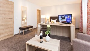 Une chambre d’hôtel avec une porte en bois, un banc, un bureau avec une télévision, un téléphone, une lampe, un vase avec une fleur et une bouteille de vin.