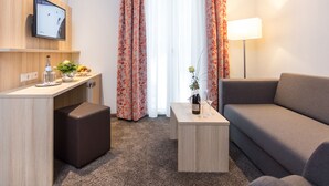Une chambre d’hôtel équipée d’une télévision à écran plat, d’un bureau agrémenté d’un vase de fleurs, d’une petite table avec une bouteille et un verre, d’un canapé et d’une lampe.