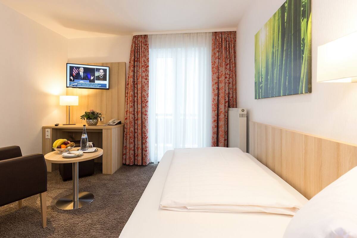 A hotel room with a bed, a desk with a computer, a chair, a small table with a fruit bowl, a lamp, and a painting of bamboo.