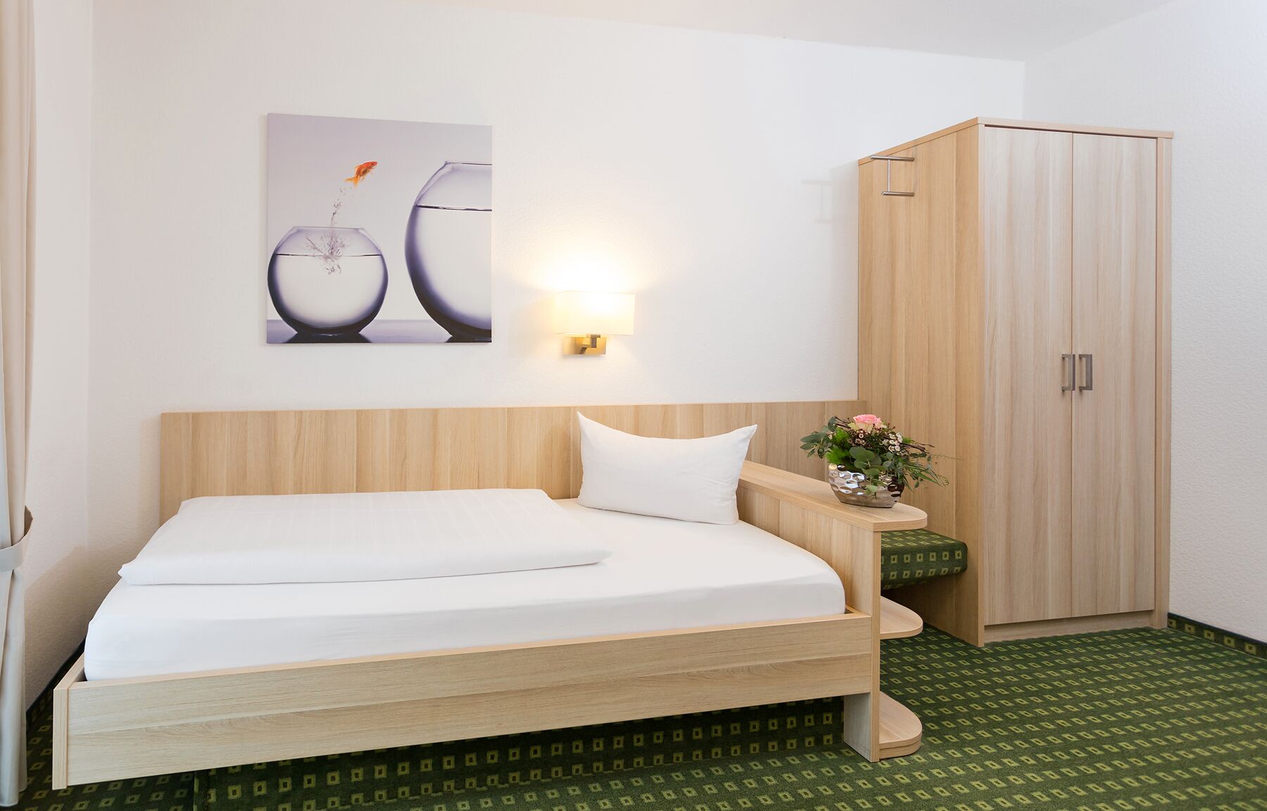 Une chambre d’hôtel comprenant un lit, une table de chevet, une lampe fixée au mur, une armoire et un tableau encadré accroché au mur.