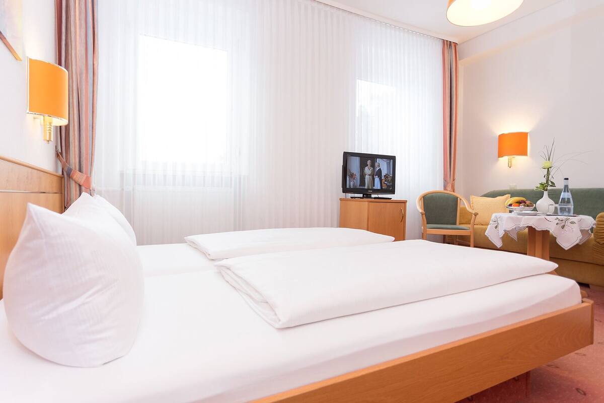 A hotel room with a bed, a television, a chair, a small table with a vase and a bottle, and a window with curtains.
