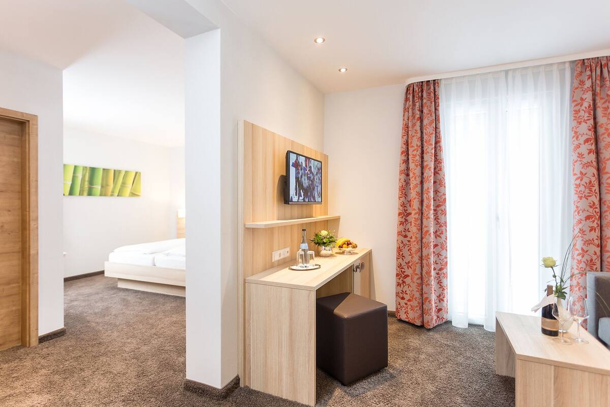 A hotel room with a bed, a desk with a computer, a small table with a vase and a bottle, and a large window with curtains.