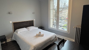 A neatly made bed in a room with a large window, a small bedside table, and a chair. - ABC Hôtel (Toul)